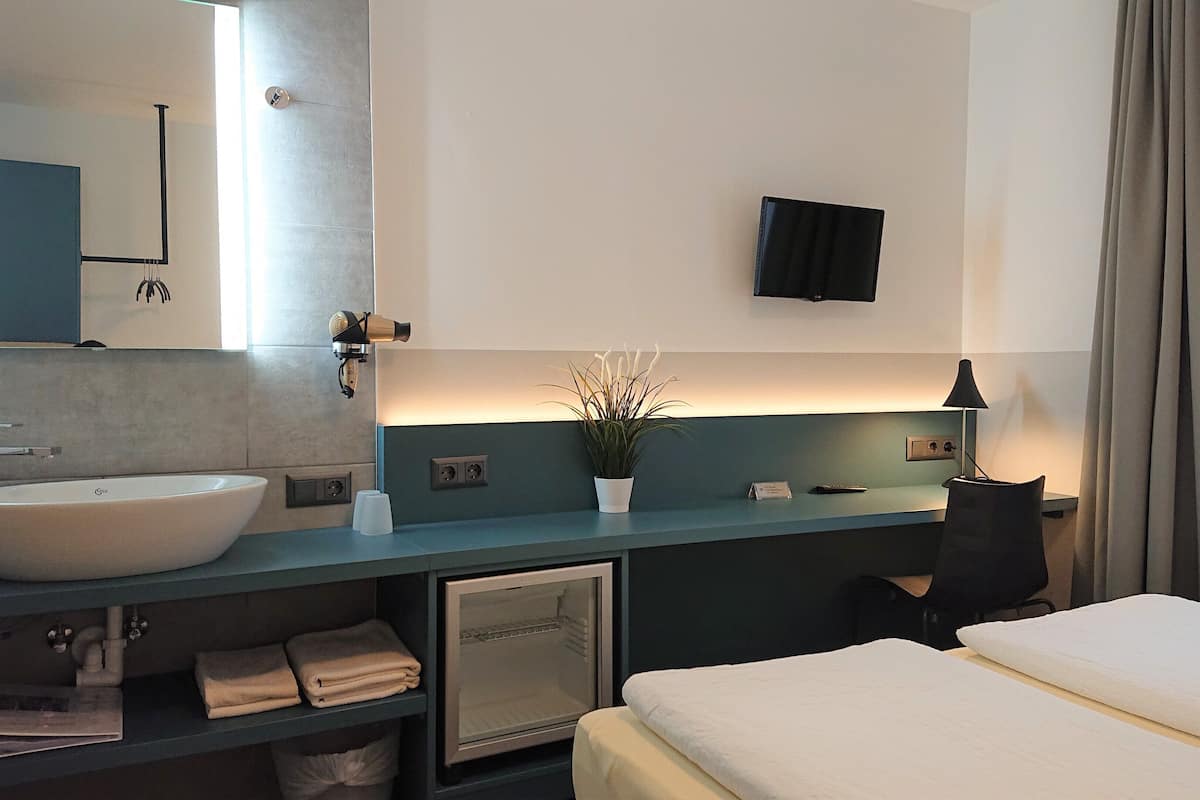 Moderni hotellihuone, jossa on suuri valkoinen pesuallas, peili, pieni ruukkukasvi, työpöytä lampun kanssa ja sänky.