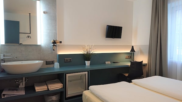 Une chambre d’hôtel moderne avec un grand lavabo blanc, un miroir, une petite plante en pot, un bureau avec une lampe et un lit.