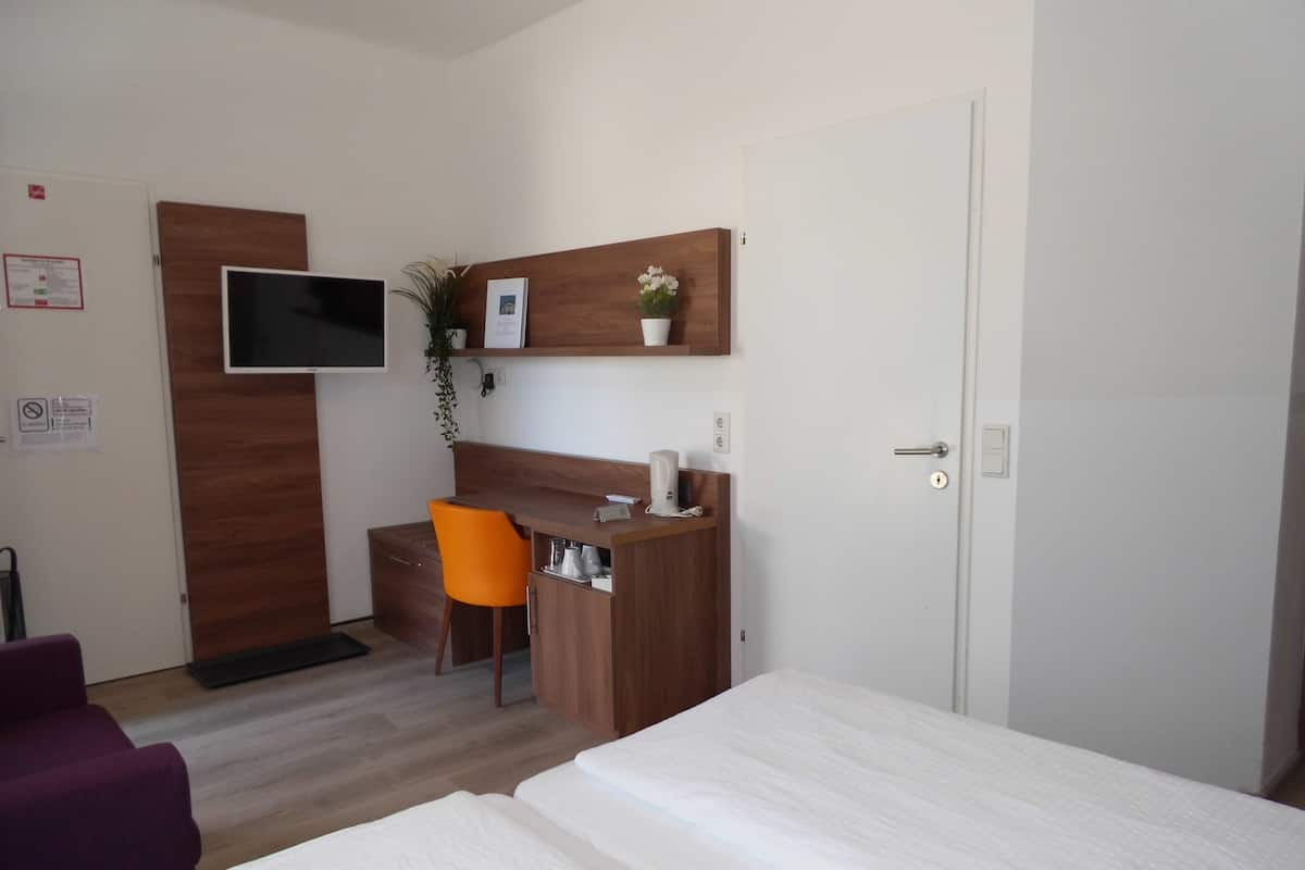 Hotellihuoneessa on sänky, työpöytä tuolilla, seinälle asennettu televisio ja pieni kasvi työpöydällä.
