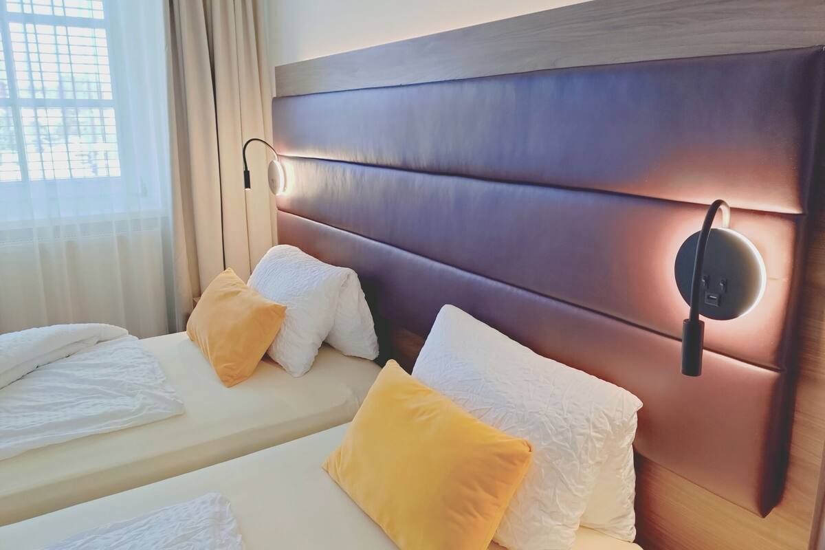 Hotellihuone, jossa on kaksi sänkyä. Jokaisessa sängyssä on valkoiset ja keltaiset tyynyt, puinen pääty ja seinään kiinnitetty valaisin.
