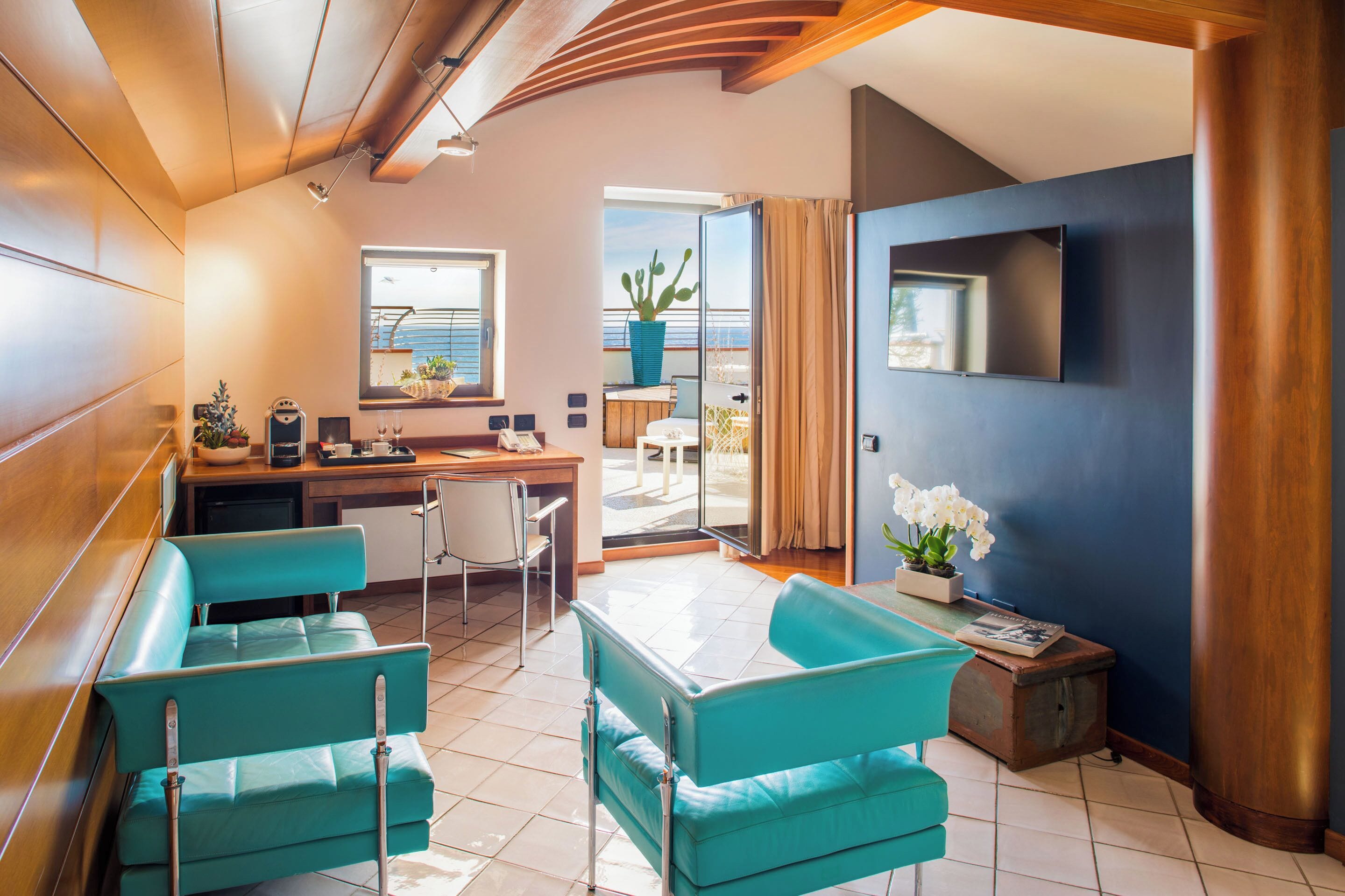 A modern hotel room with teal furniture, a desk, a chair, a small table, and a potted plant.