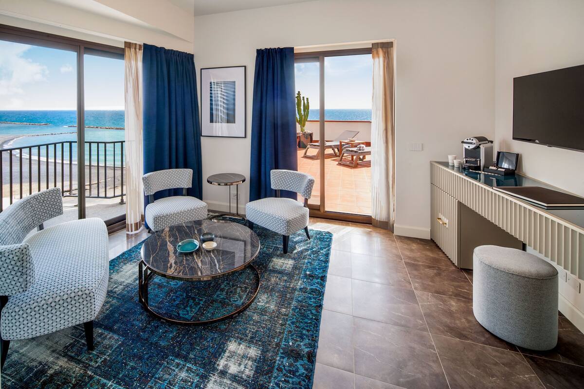 A modern hotel room with a balcony, a round glass coffee table, patterned armchairs, and a kitchen area with a flat-screen TV.