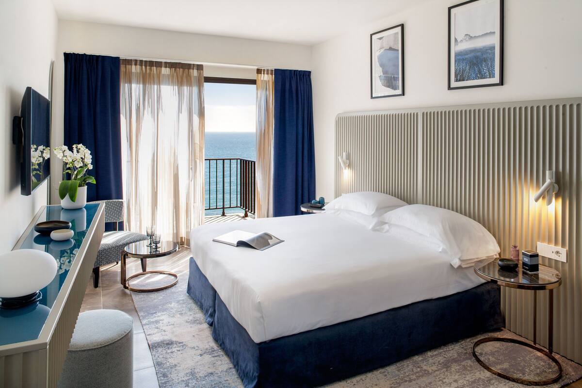 A hotel room with a bed, a desk with a chair, a small table, a mirror, and a view of the sea through large windows.