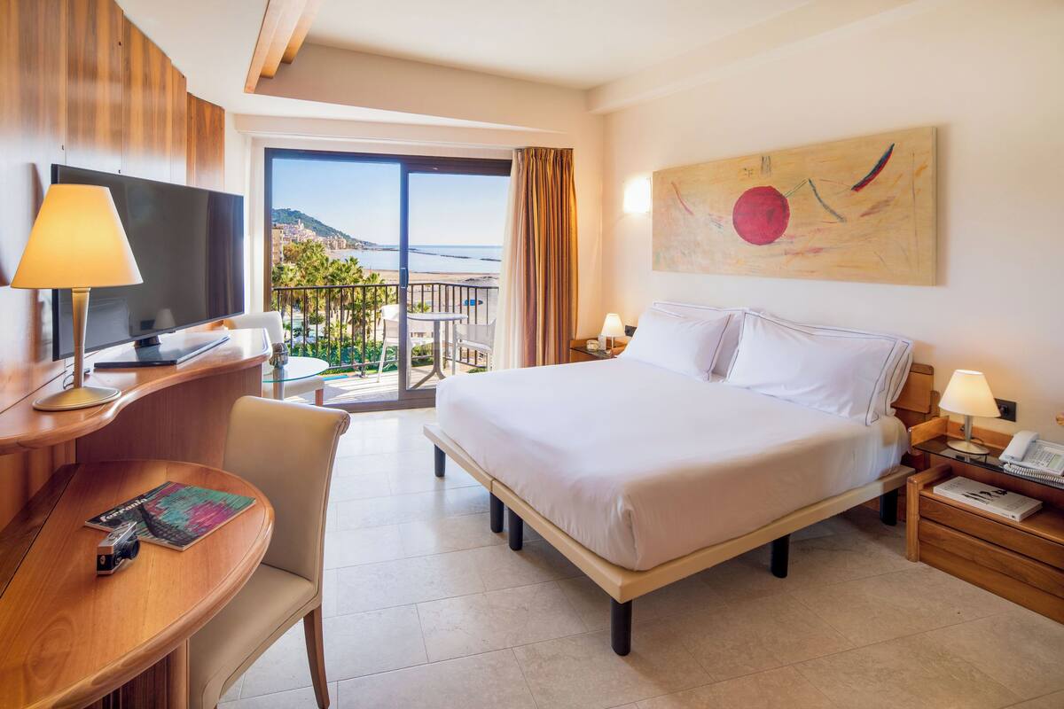 A hotel room with a bed, desk, chair, and a large window offering a view of the sea.