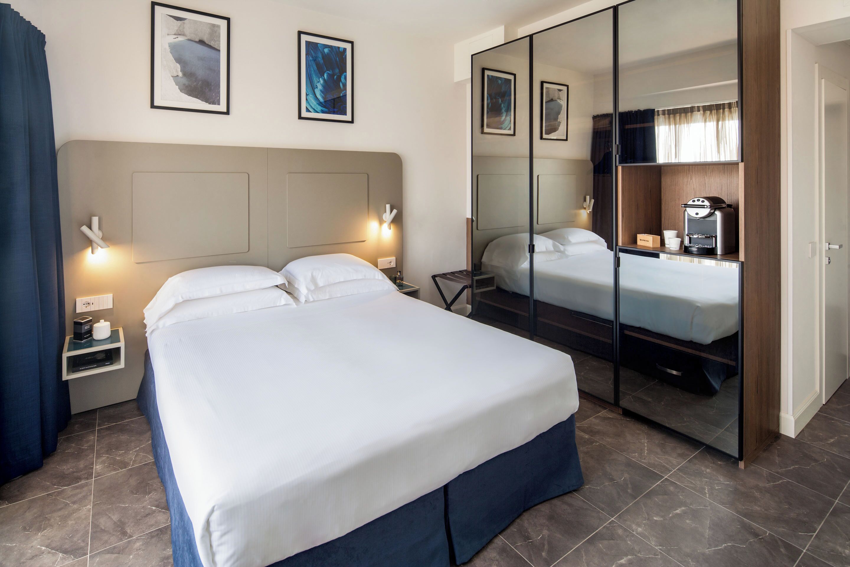A hotel room with a large bed, bedside tables, a wardrobe with a mirror, and artwork on the walls.