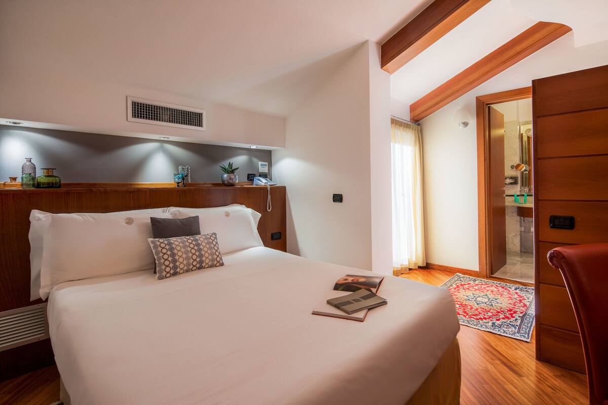 A hotel room with a large bed, a wooden headboard, a bedside table with a lamp, and a red carpet leading to a bathroom.