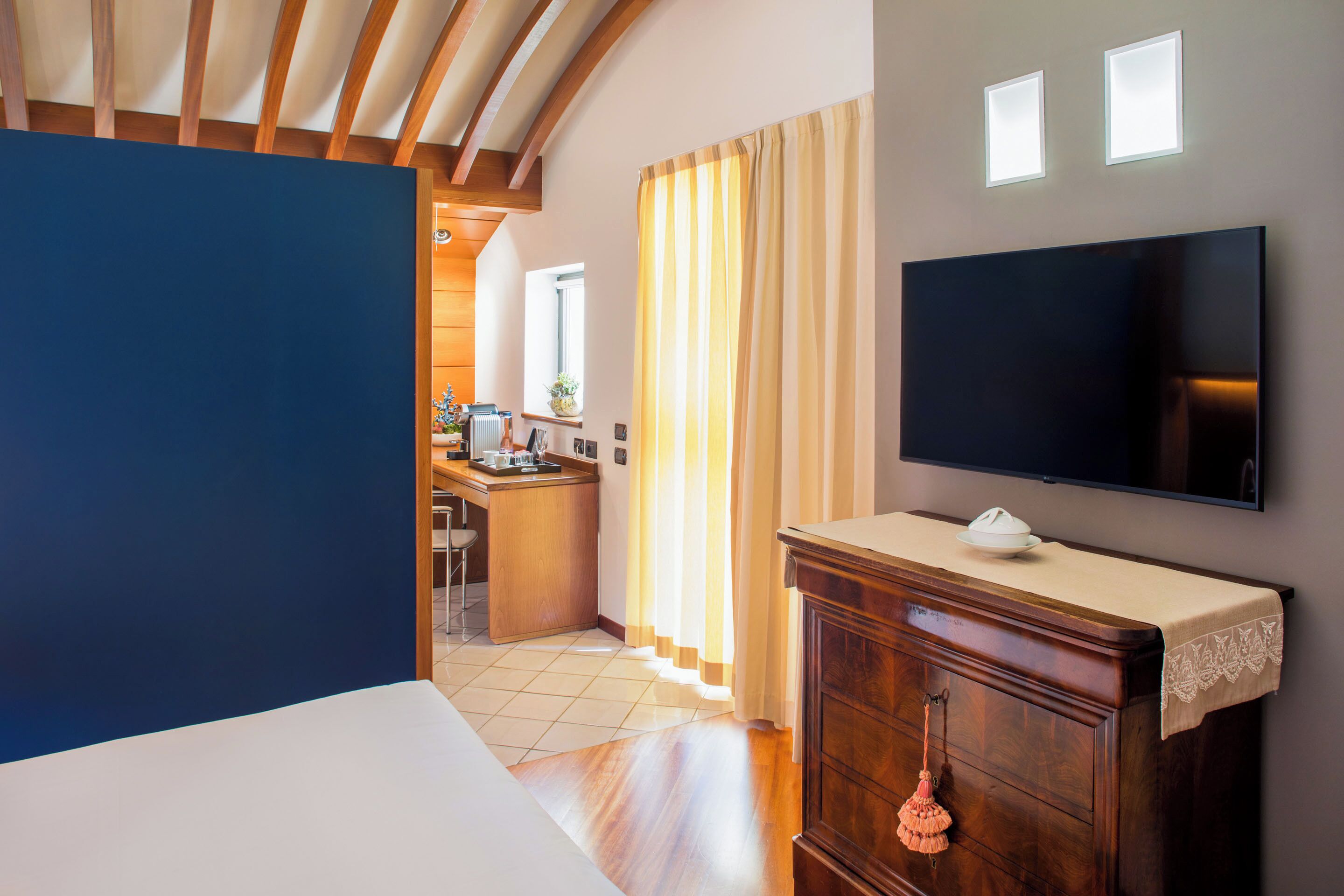 A hotel room with a large bed, a wooden dresser, a flat-screen TV, and a kitchen area in the background.