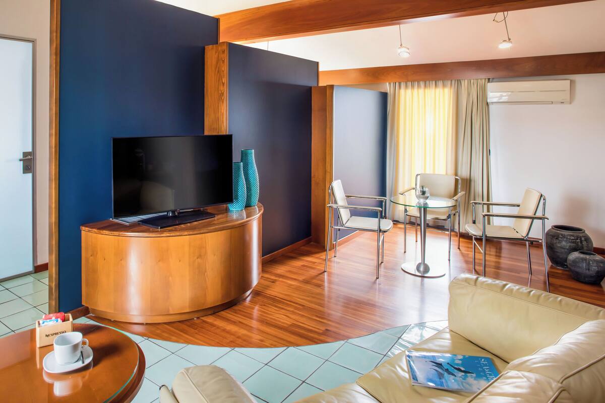 A modern hotel room with a wooden reception desk, a flat-screen TV, a sofa, a small table with a cup, and a dining area with chairs.