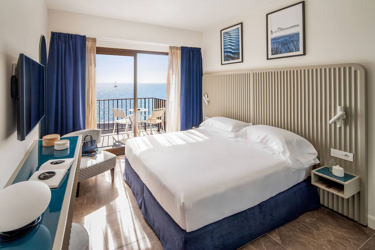 A hotel room with a large bed, a TV, a desk with a chair, a small table, and a view of the sea through the balcony doors.