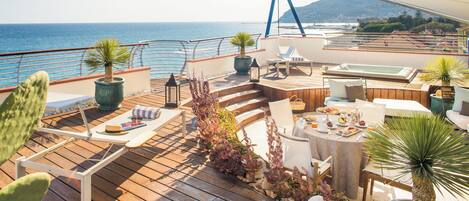 A sunlit deck with wooden flooring, potted plants, outdoor seating, and a view of the sea.