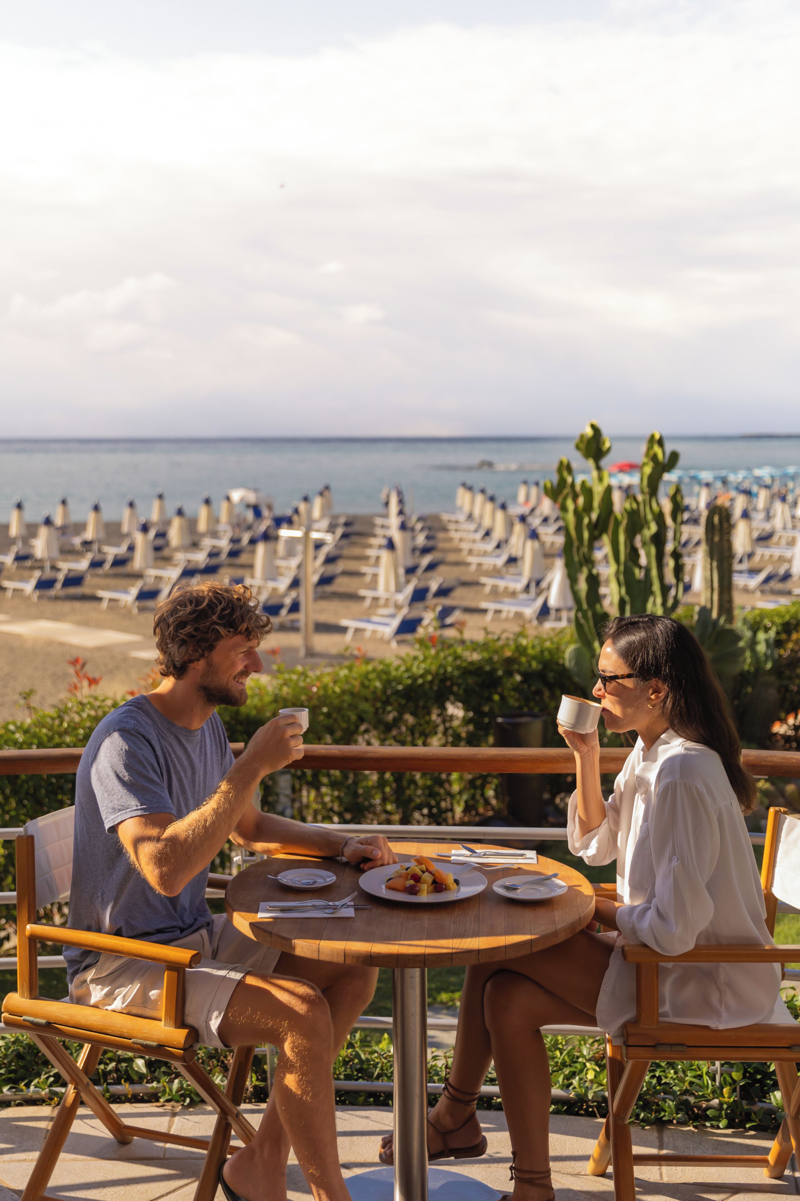 Two people seated at a round table with a view of the beach and cacti in the background.