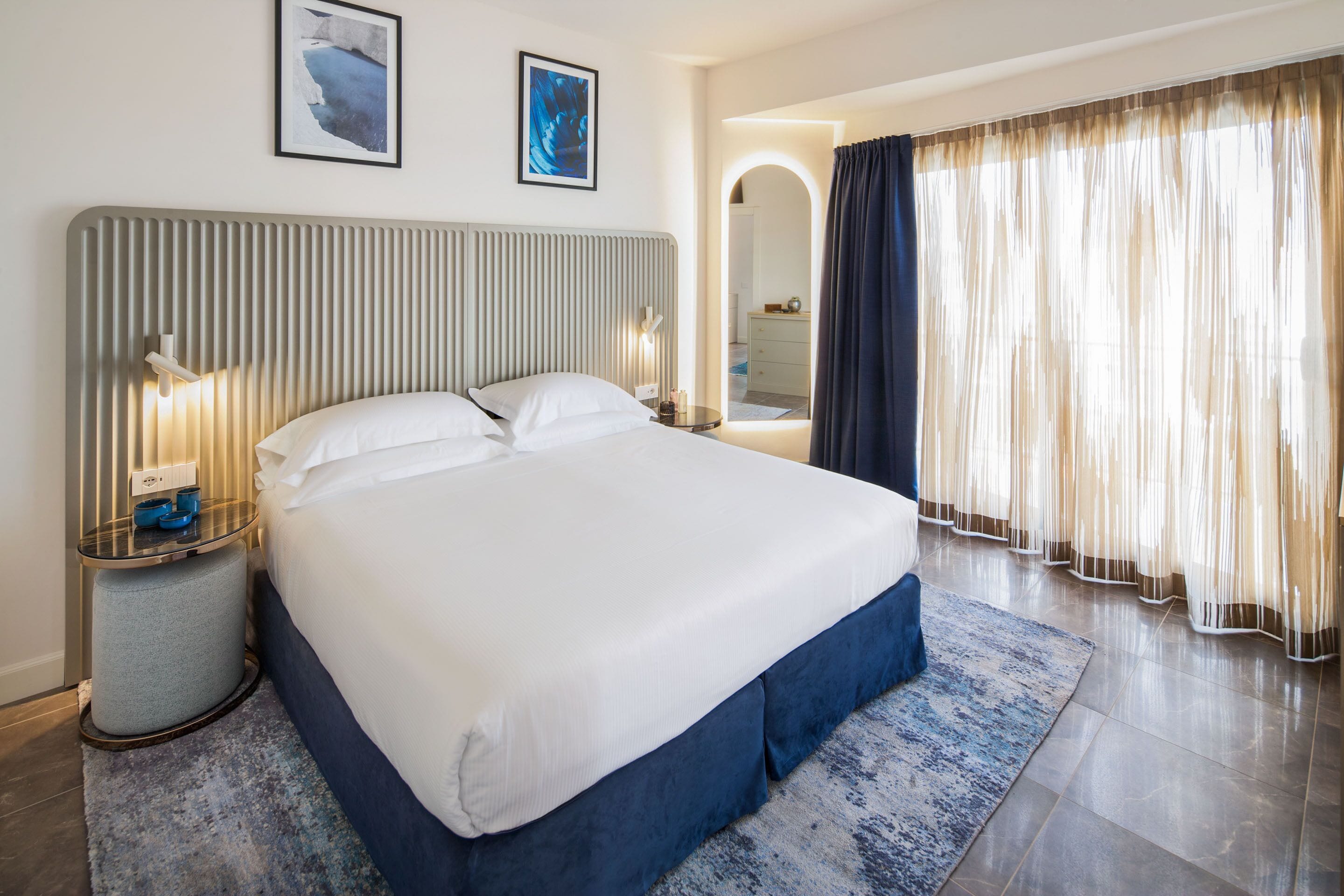 Une chambre d’hôtel moderne avec un grand lit, des lampes de chevet, une petite table ronde et une vue sur la salle de bain à travers une porte en arc de cercle.