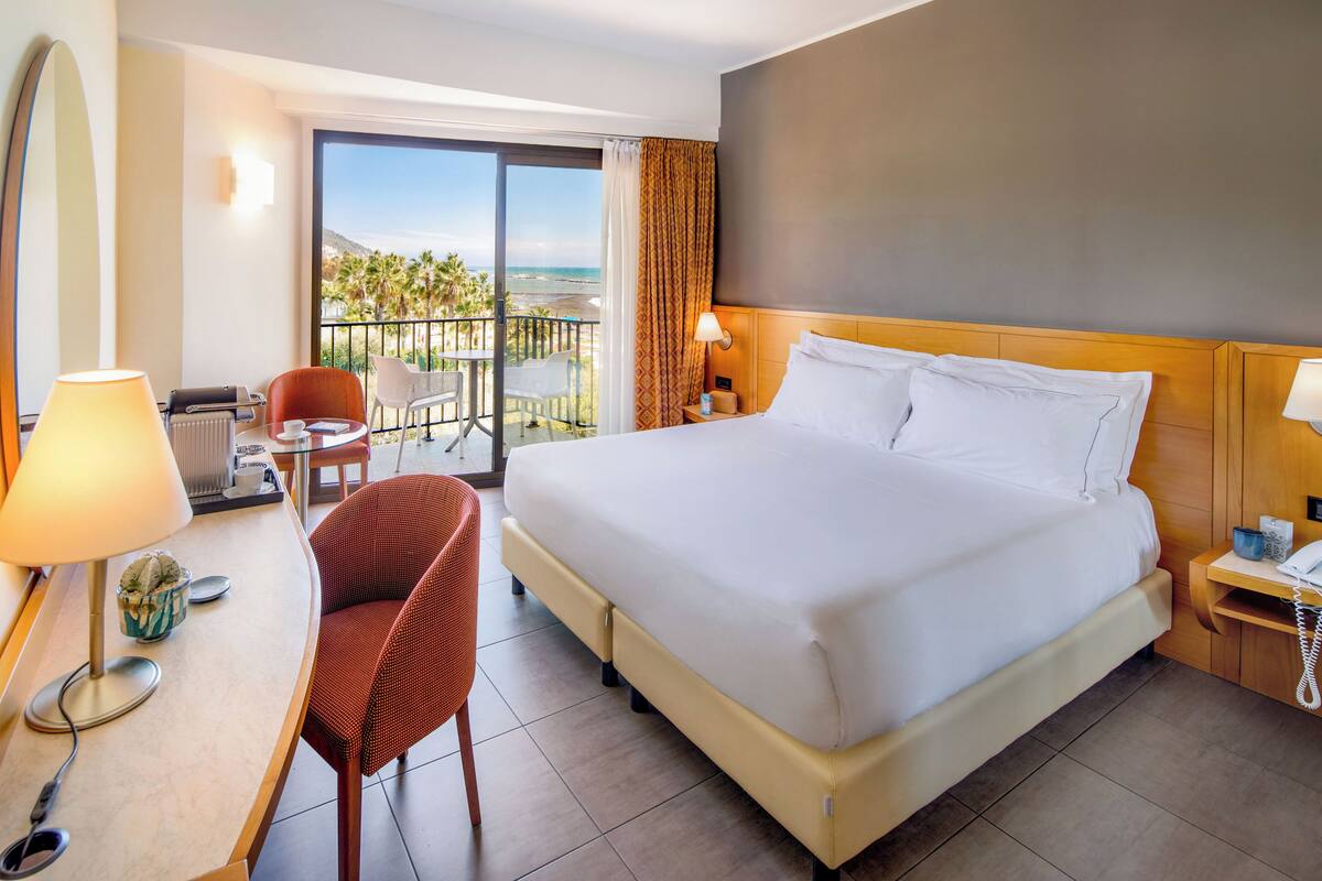 A hotel room with a large bed, a desk with a lamp, a chair, and a view of the beach.
