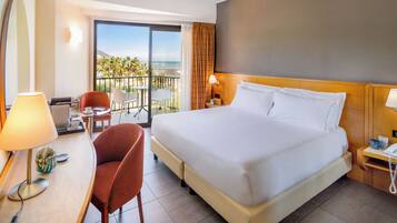 Habitación de hotel con una cama grande, un escritorio con lámpara, una silla y vista a la playa.