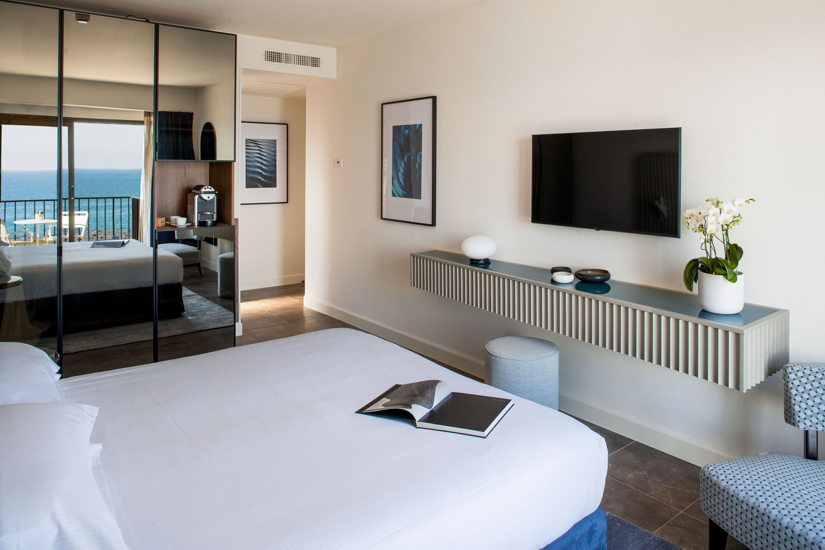 A modern hotel room with a large bed, a TV, a bedside table, a small seating area, and a view of the sea.