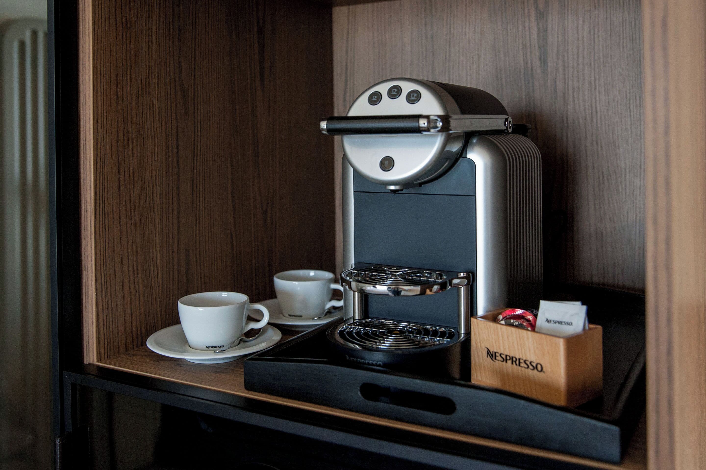 A compact espresso machine with two cups on a tray, a Nespresso coffee pod box, and a wooden panel background.