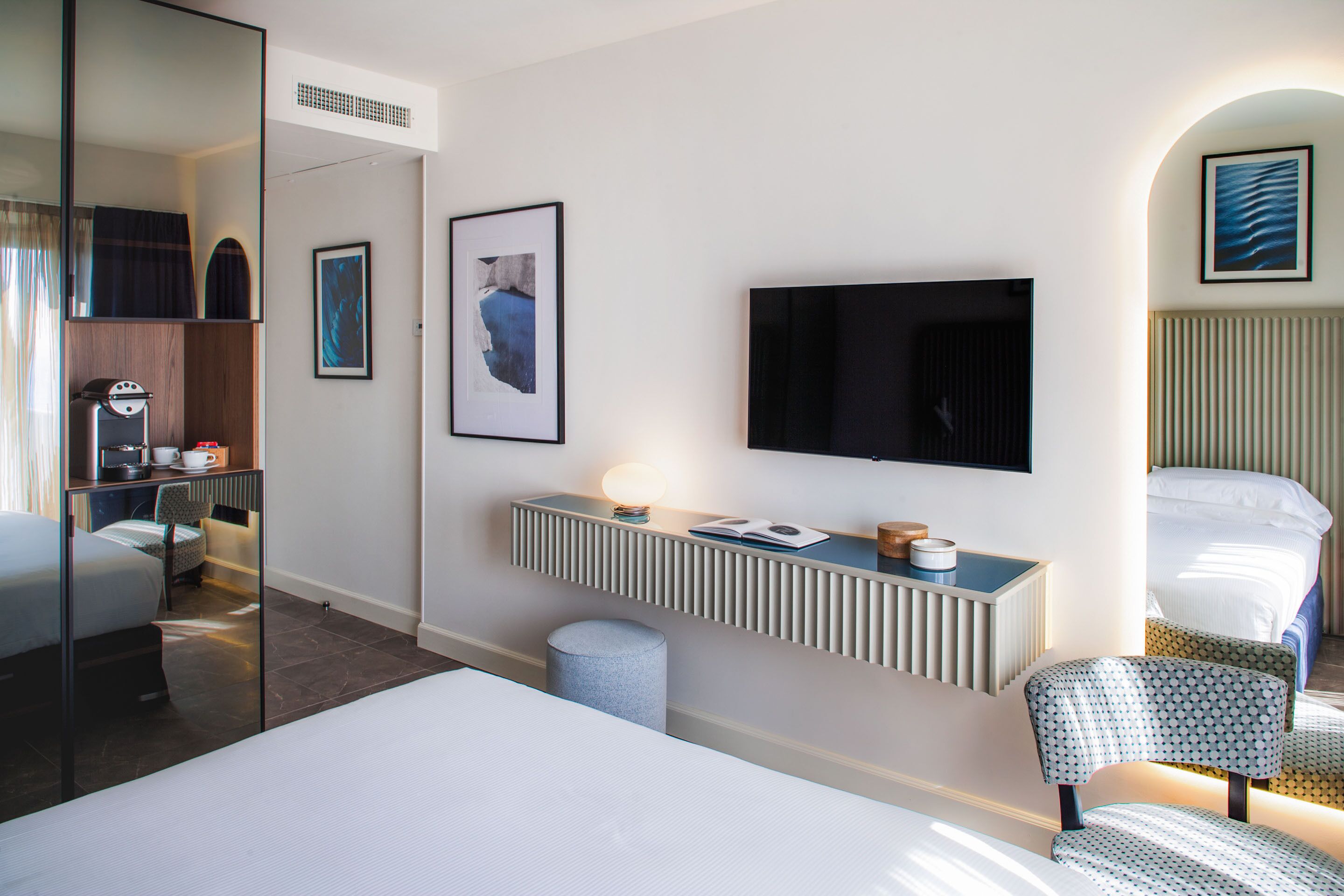 Habitación de hotel con cama, escritorio con televisor, silla, taburete pequeño, espejo y obras de arte en las paredes.