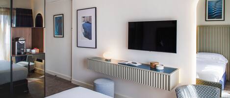 Habitación de hotel con cama, escritorio con televisor, silla, taburete pequeño, espejo y obras de arte en las paredes.