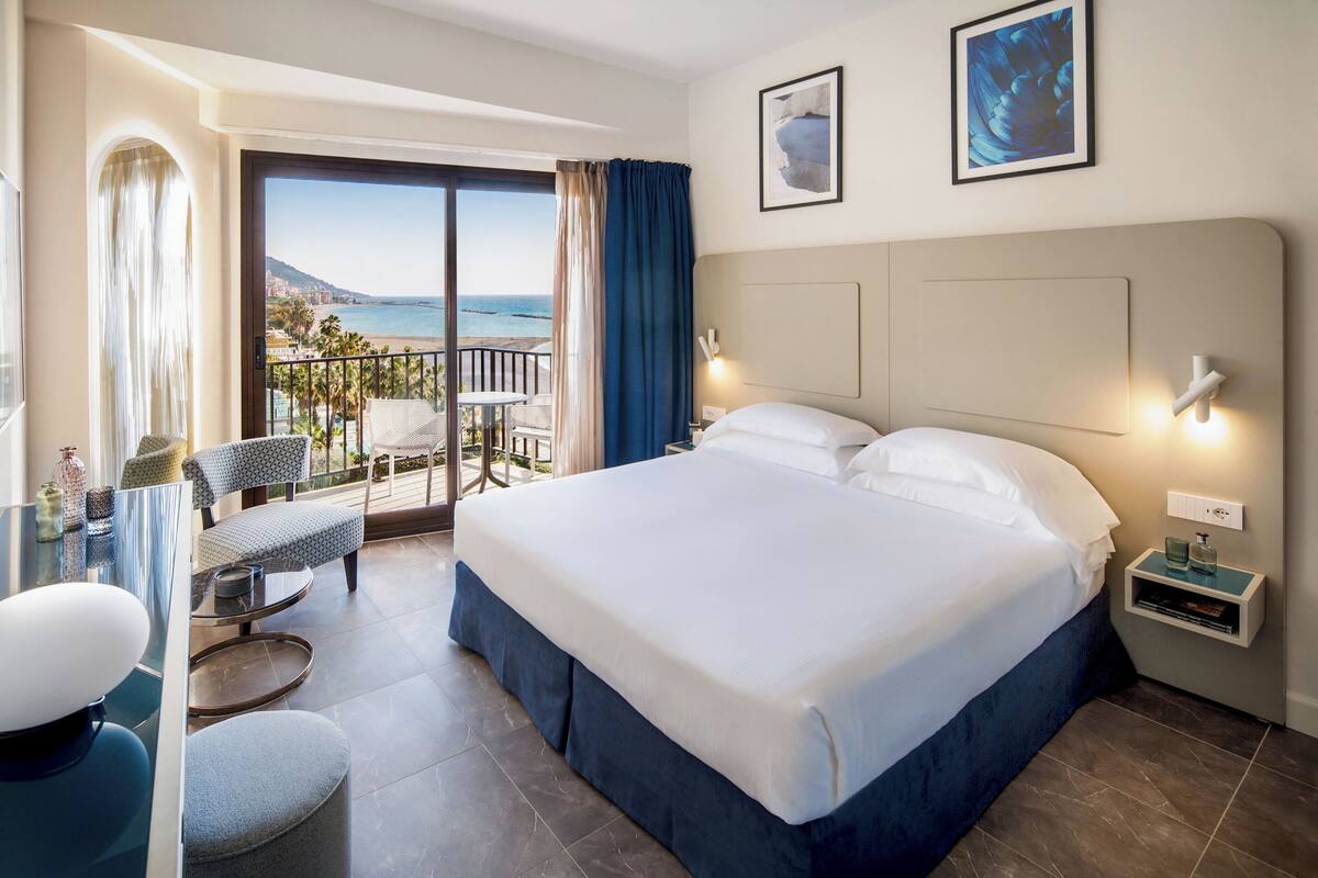 A hotel room with a large bed, a desk with a chair, a balcony with a view of the sea, and two framed pictures on the wall.