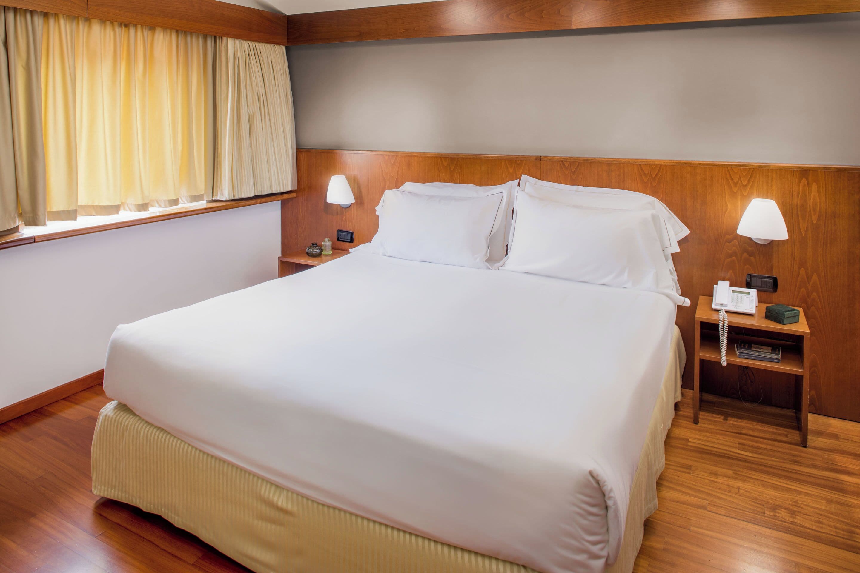 Una cama grande con ropa blanca de cama, cabecera de madera, lámparas de noche y teléfono junto a la cama.