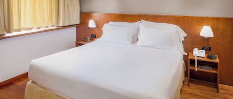 Una cama grande con ropa blanca de cama, cabecera de madera, lámparas de noche y teléfono junto a la cama.