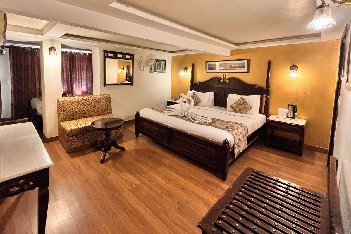 A hotel room with a wooden floor, a bed with a headboard, a bedside table, a chair, a desk, and a mirror.