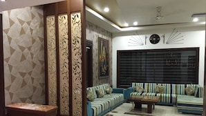 A modern living room with a sofa set, decorative panels, a clock, and a window with blinds. - Hotel Ratnawali (Jaipur)