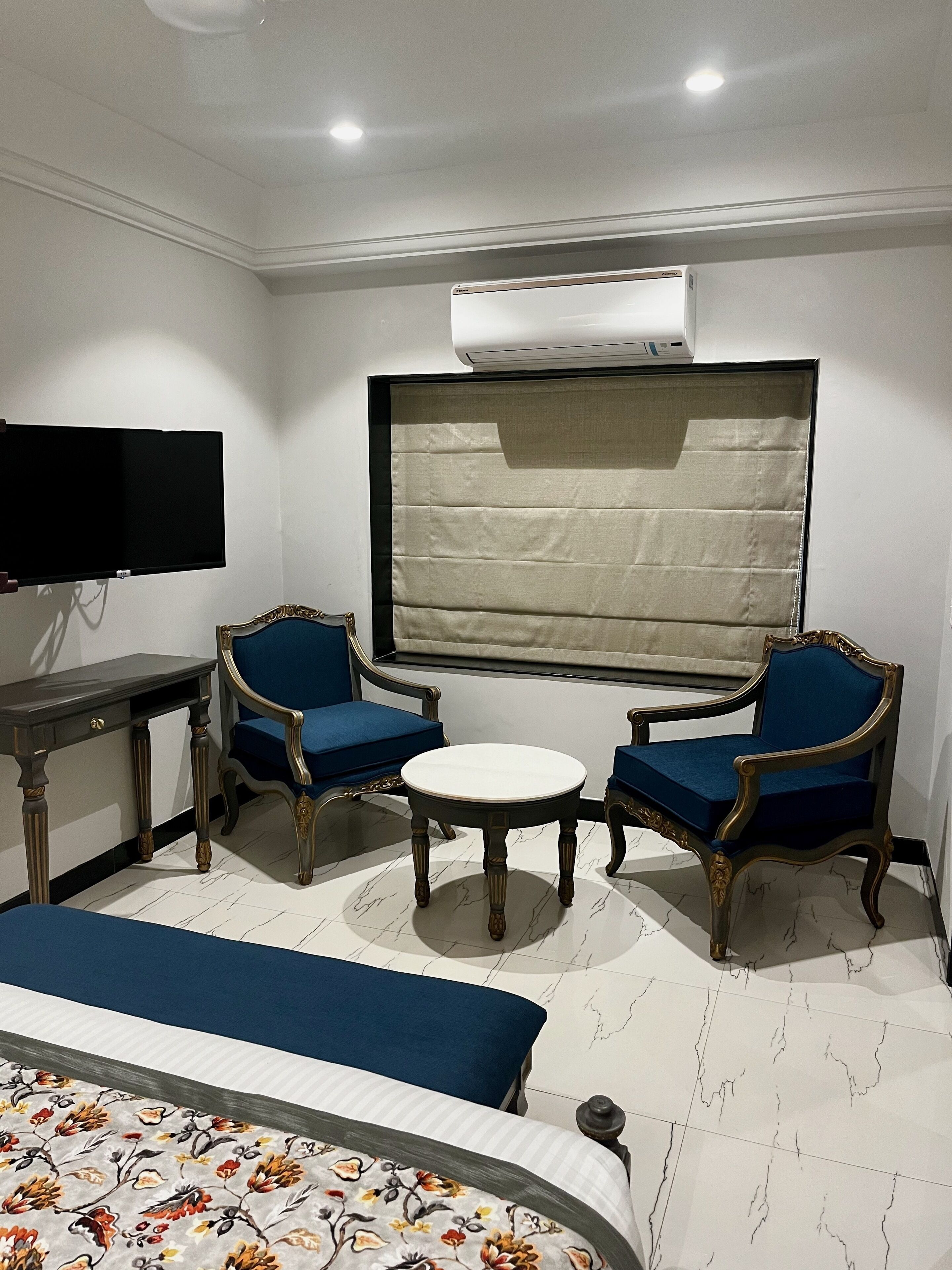 Une chambre d’hôtel avec un lit, deux fauteuils bleus, une petite table blanche, un téléviseur fixé au mur et un appareil de climatisation placé au-dessus de la fenêtre.