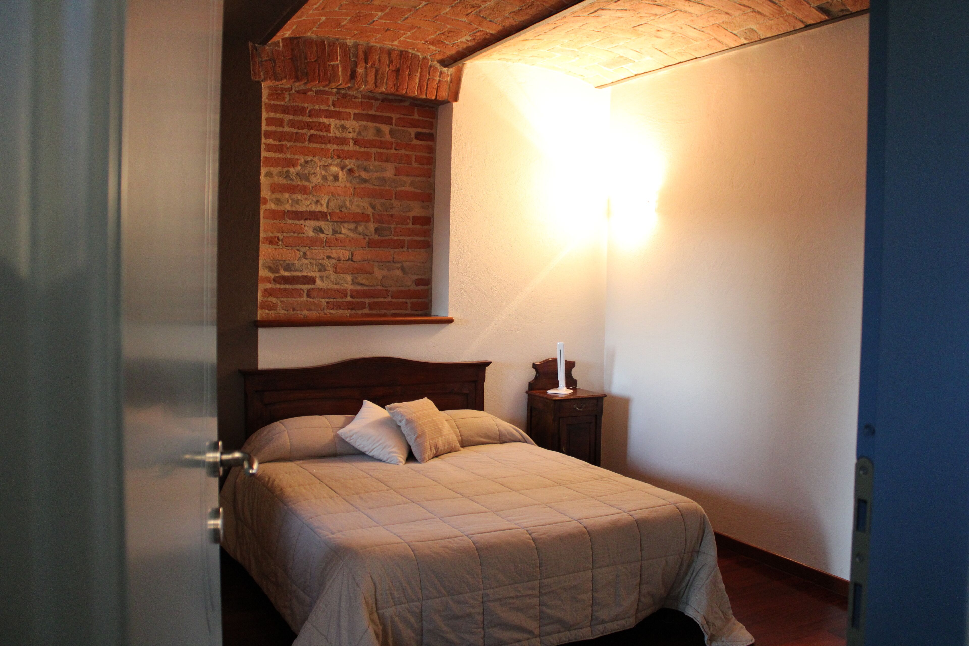 A bedroom with a brick wall, a wooden headboard, a bed with a quilt, a bedside table with a lamp, and a door.