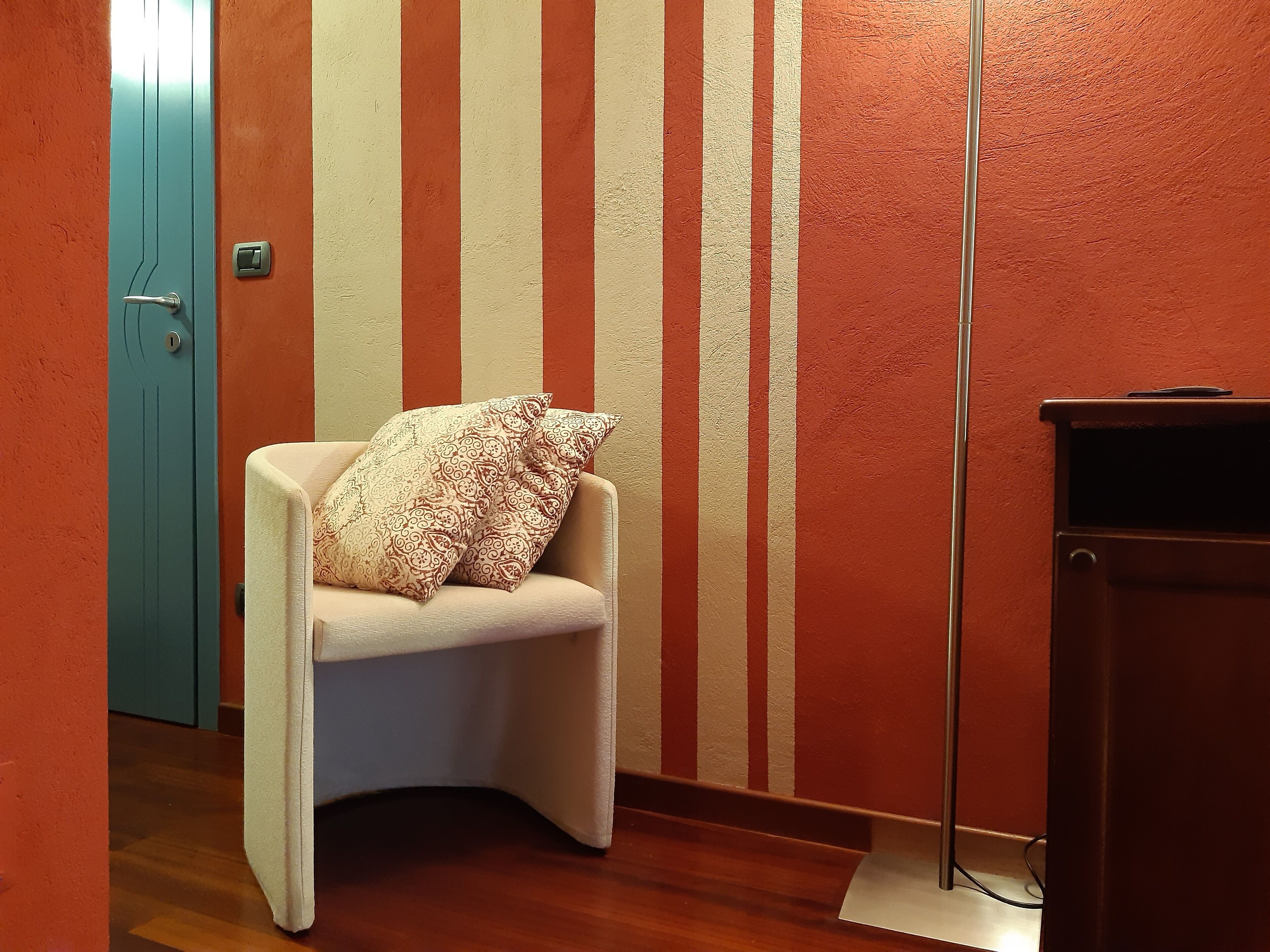 A corner of a room with a teal door, a white armchair with patterned cushions, and a wall with vertical stripes.