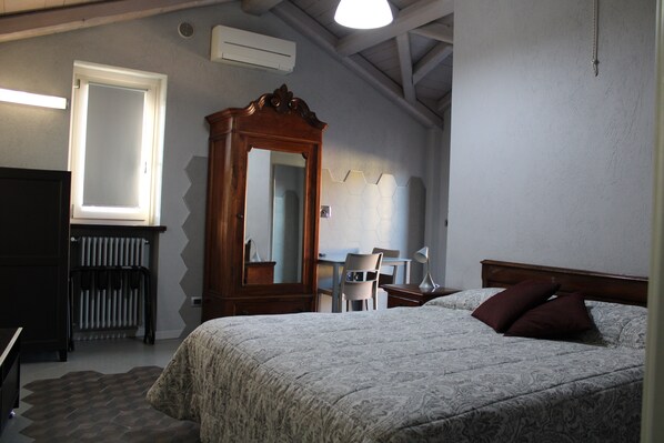 A bedroom with a bed, a wooden wardrobe, a desk with a chair, a radiator, and a large mirror.