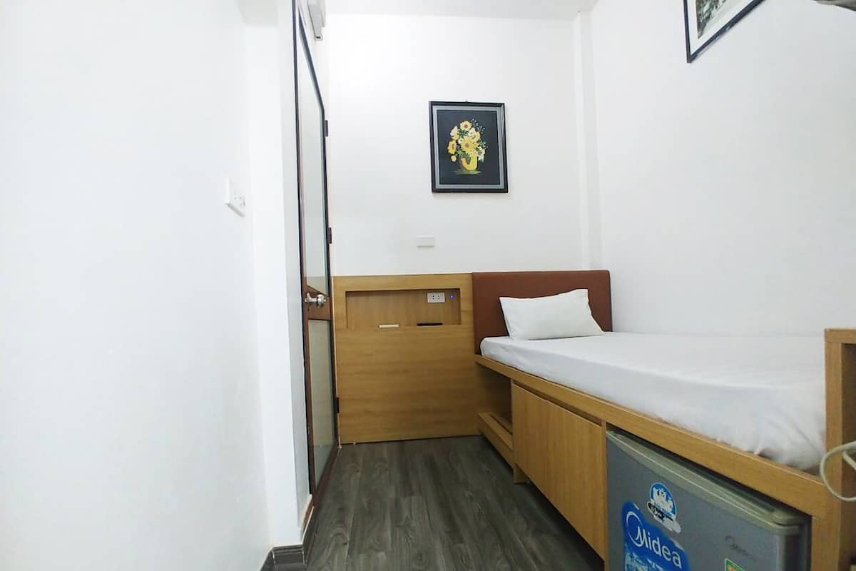 A small, compact room with a single bed, a wooden wardrobe, and a small kitchenette area with a fridge.