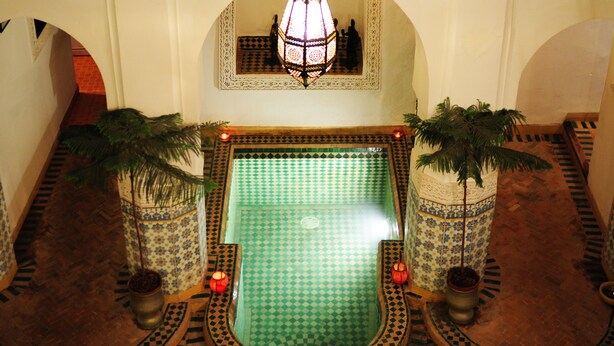 Una piscina con diseños geométricos de azulejos, rodeada de columnas ornamentales y palmeras en macetas.