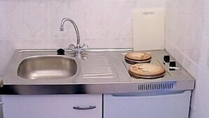 Een kleine keuken met een RVS spoelbak, één kastje en een klein aanrecht met een broodrooster en een bord.