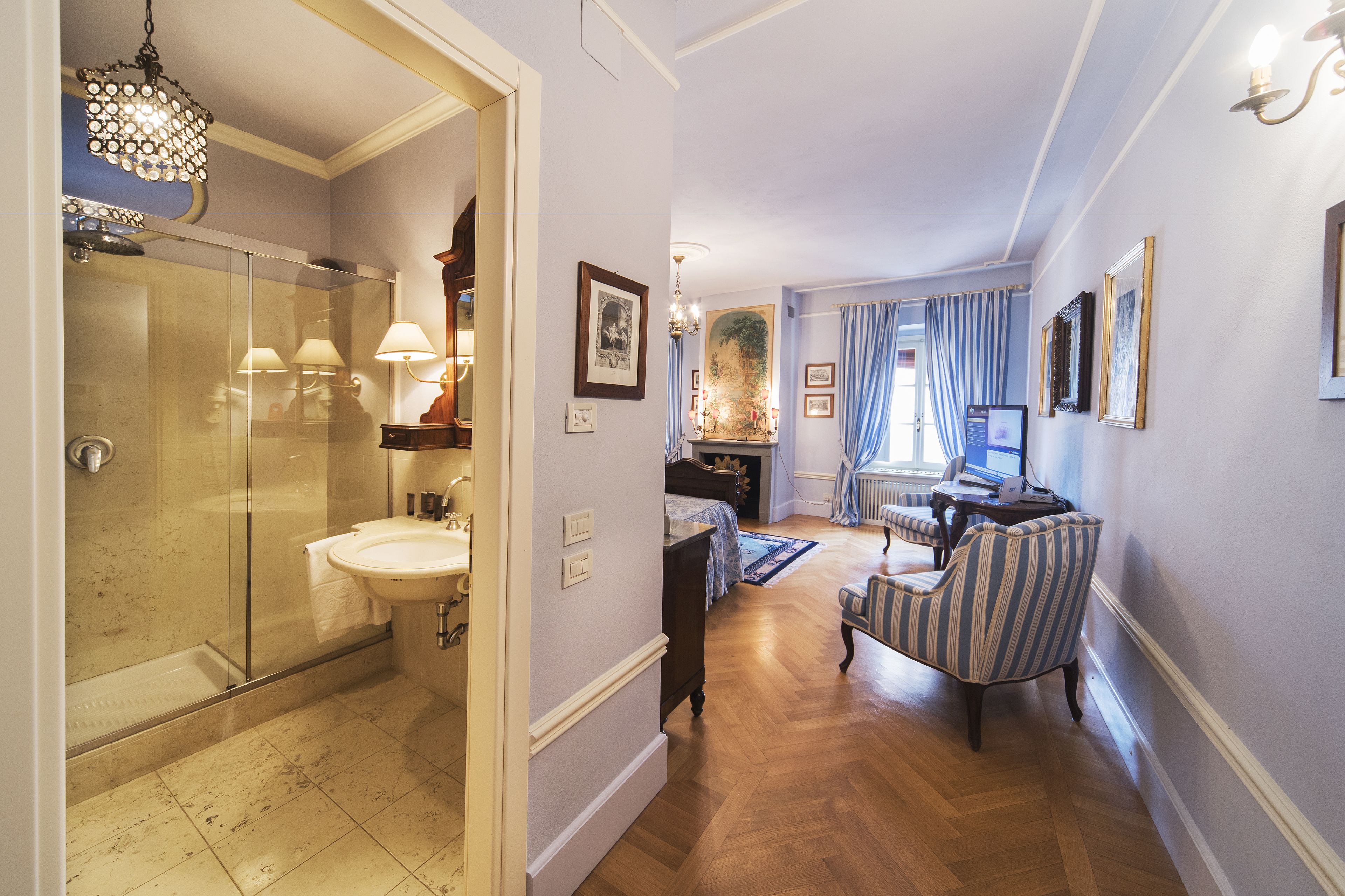 A hotel room with a glass shower, a sink, a bed, a desk, and a chair.