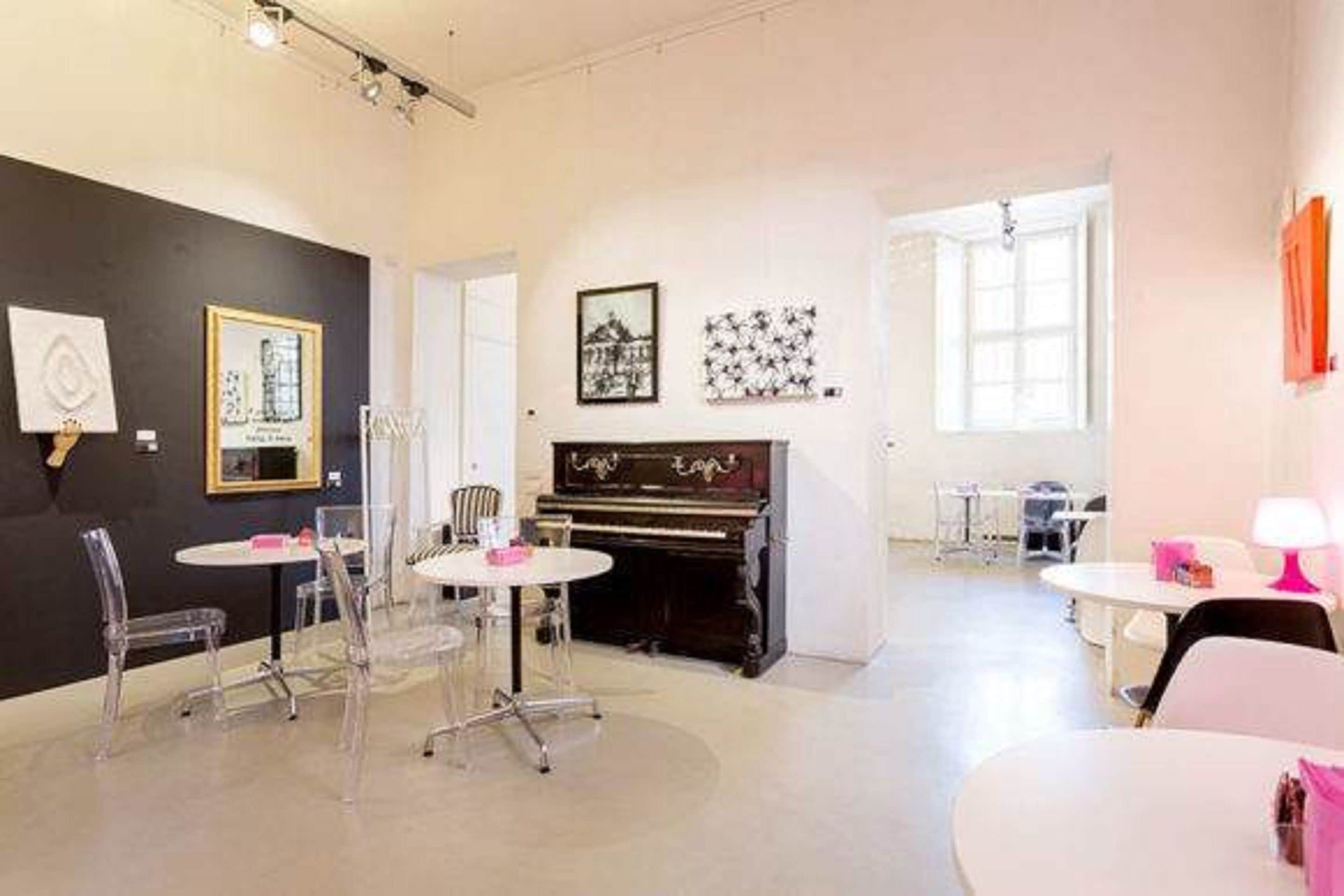 A room with a piano, clear chairs, and tables. There are framed pictures on the wall.