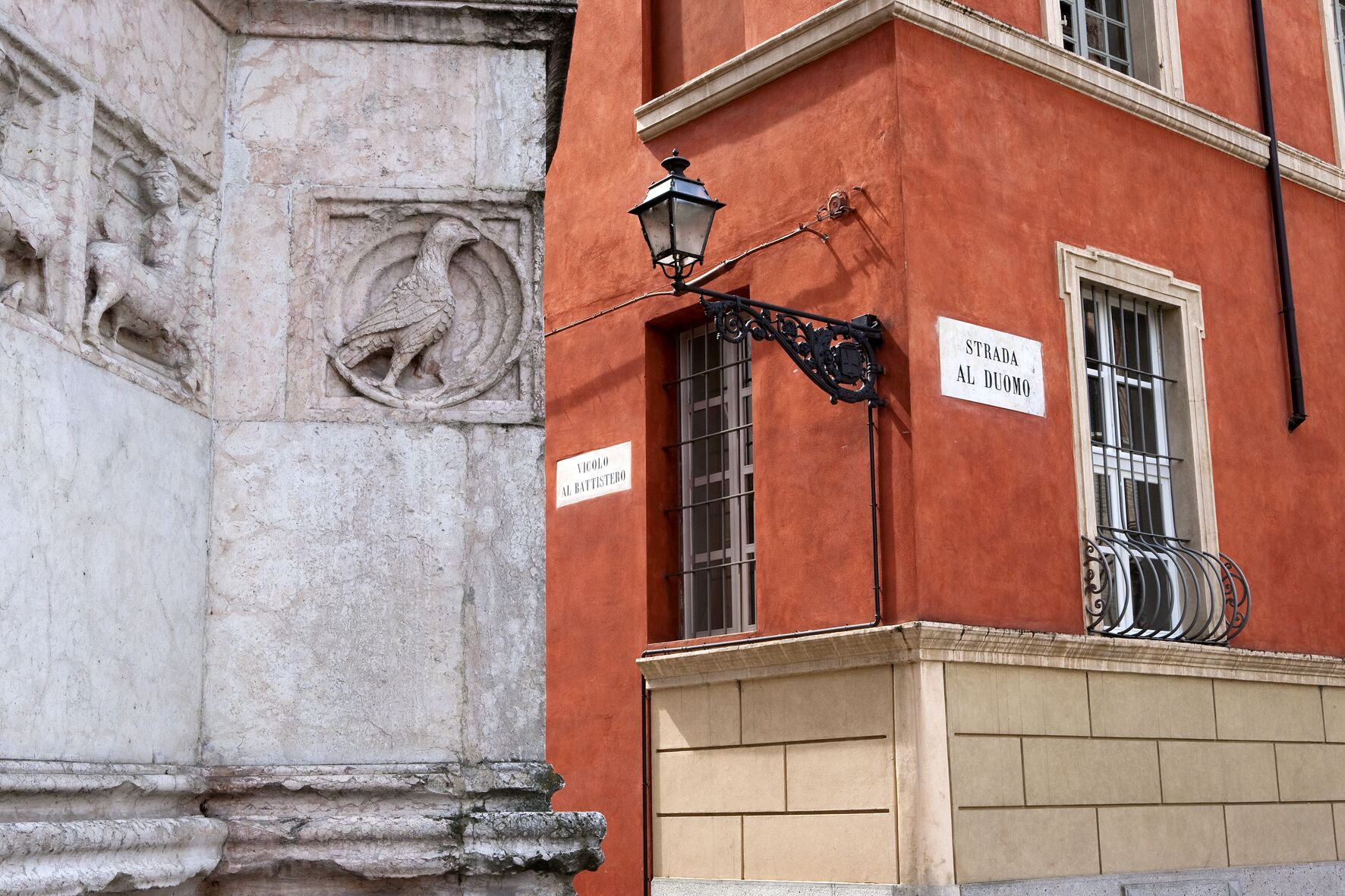 A building facade with a stone relief, a street lamp, and signs indicating 'Vicolo Al Battistero' and 'Strada Al Duomo'.