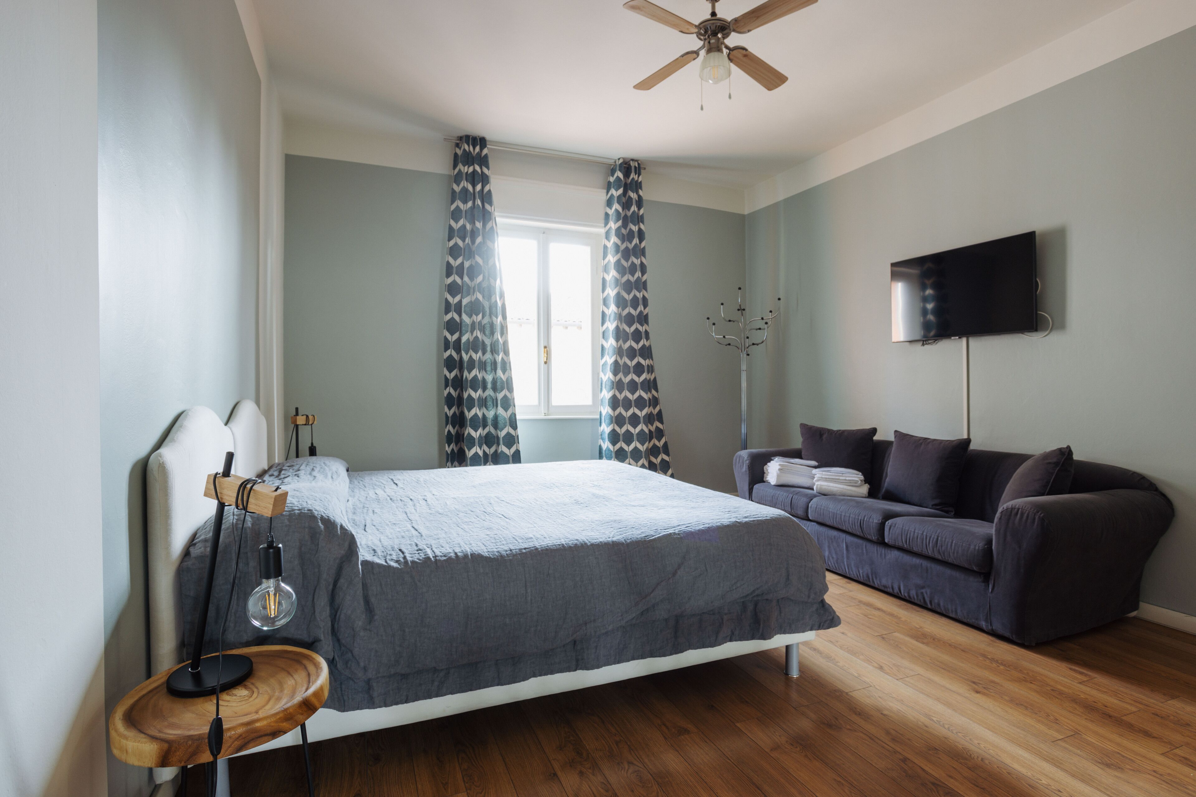 A bedroom with a bed, a wooden floor, a ceiling fan, a TV, and a window with curtains.