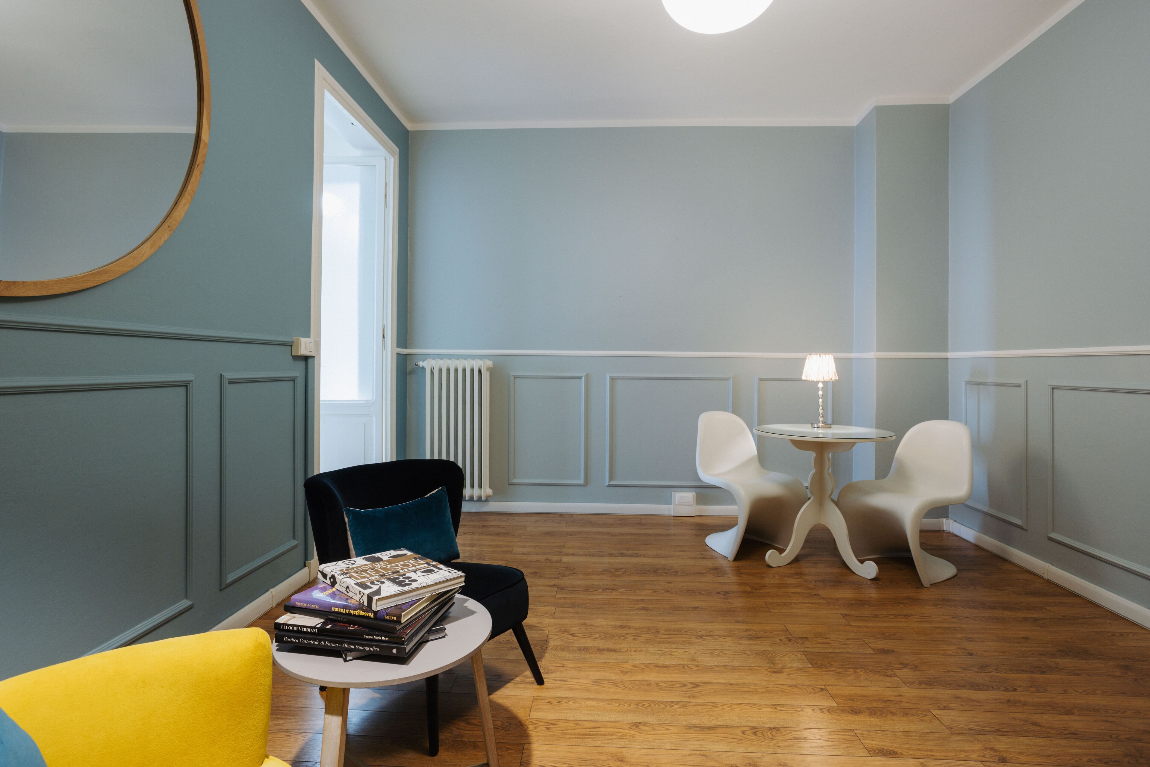 Una habitación con piso de madera, un espejo redondo, una pequeña mesa con libros y dos sillas blancas.