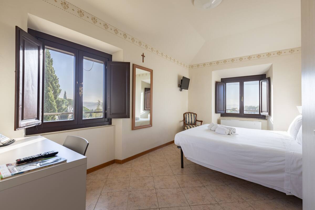 Une chambre d’hôtel équipée d’un lit, d’un bureau, d’une chaise, d’une télévision et d’une fenêtre donnant sur des arbres.