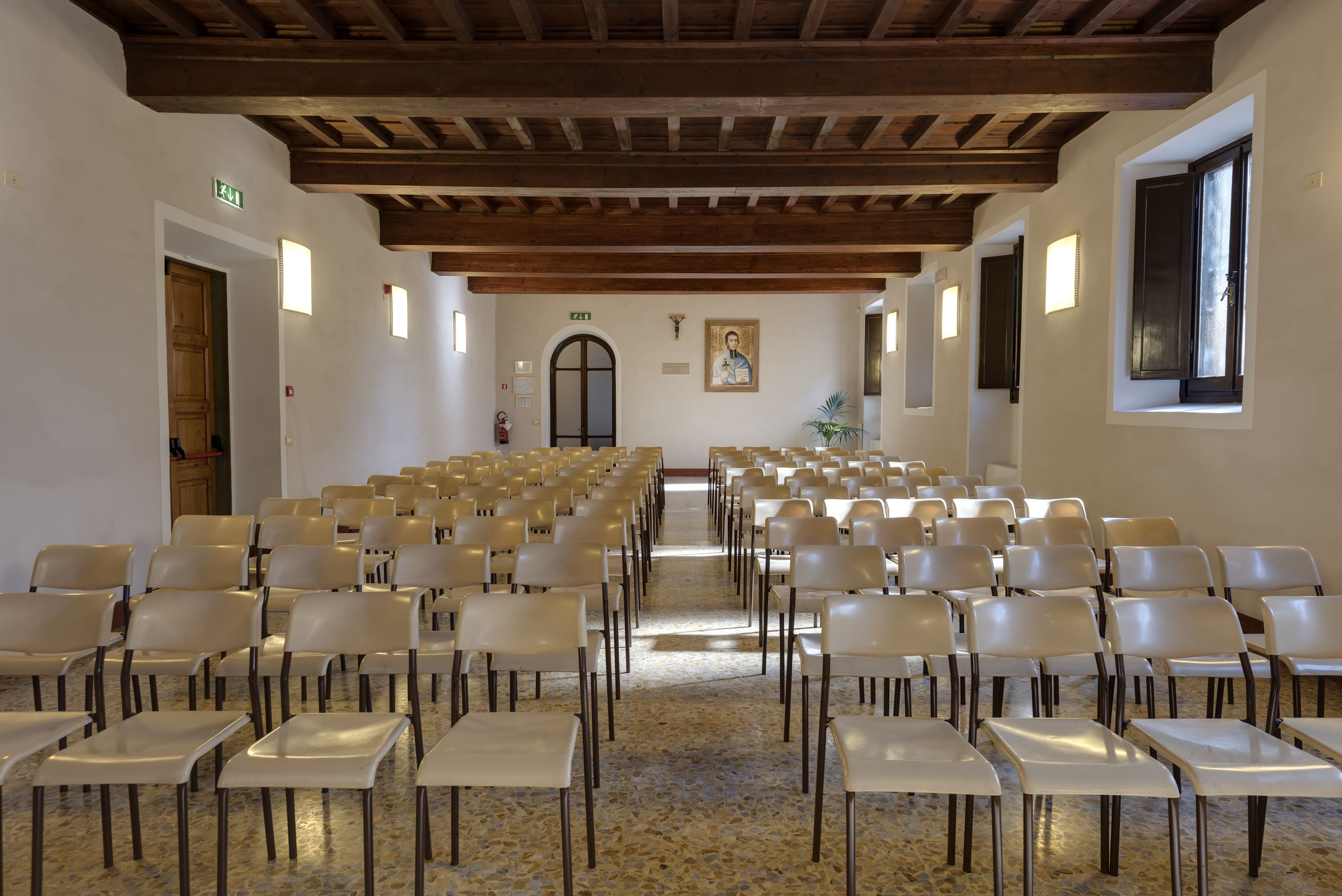 A church interior with rows of chairs and pews, wooden beams on the ceiling, and a religious painting on the wall.