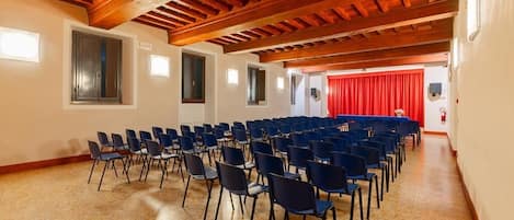 Uma sala de conferências com fileiras de cadeiras, vigas de madeira no teto e uma cortina vermelha ao fundo.
