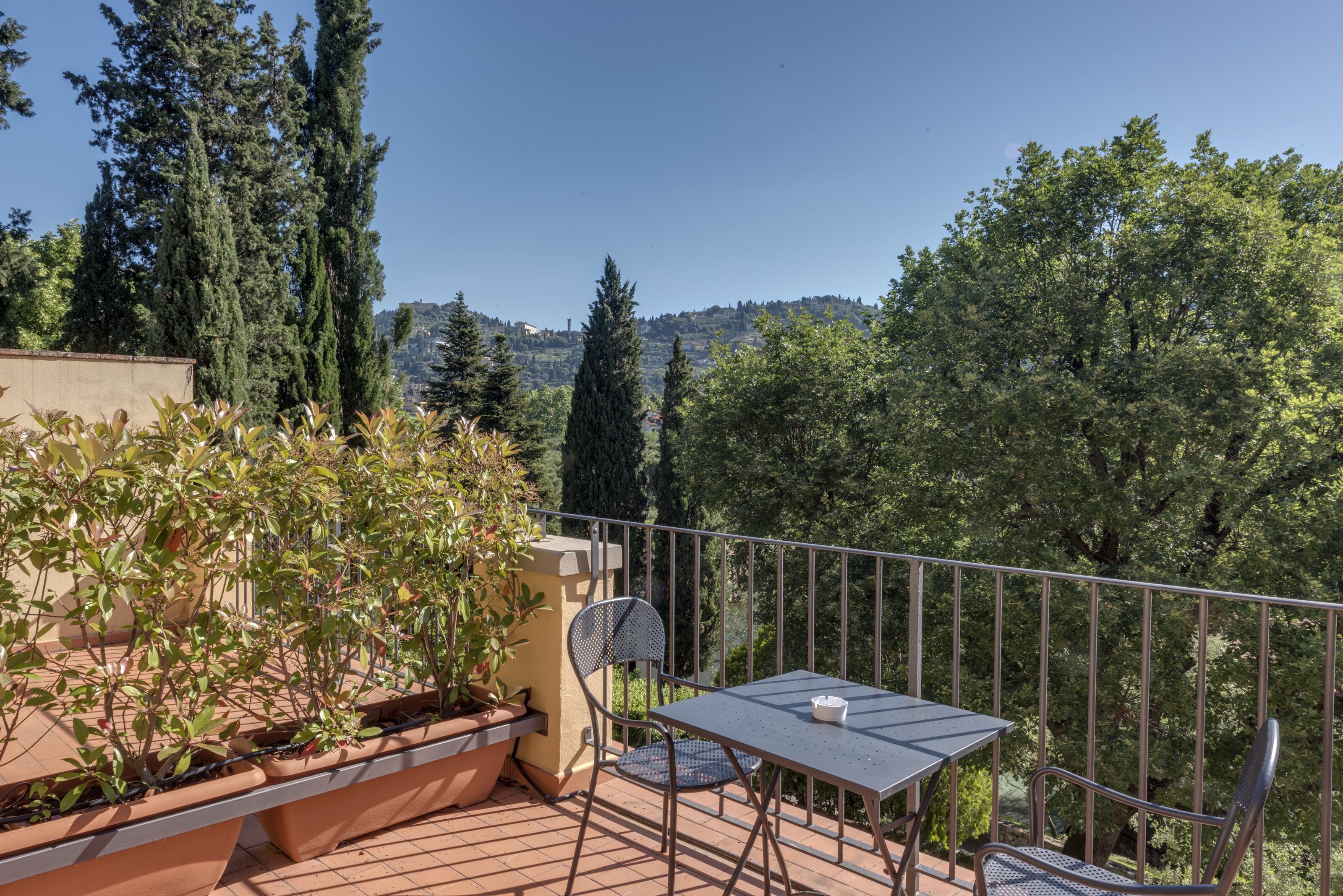 A balcony with a table and chairs, surrounded by potted plants and overlooking a scenic view of trees and hills.