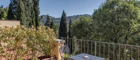 A balcony with a table and chairs, surrounded by potted plants and overlooking a scenic view of trees and hills.