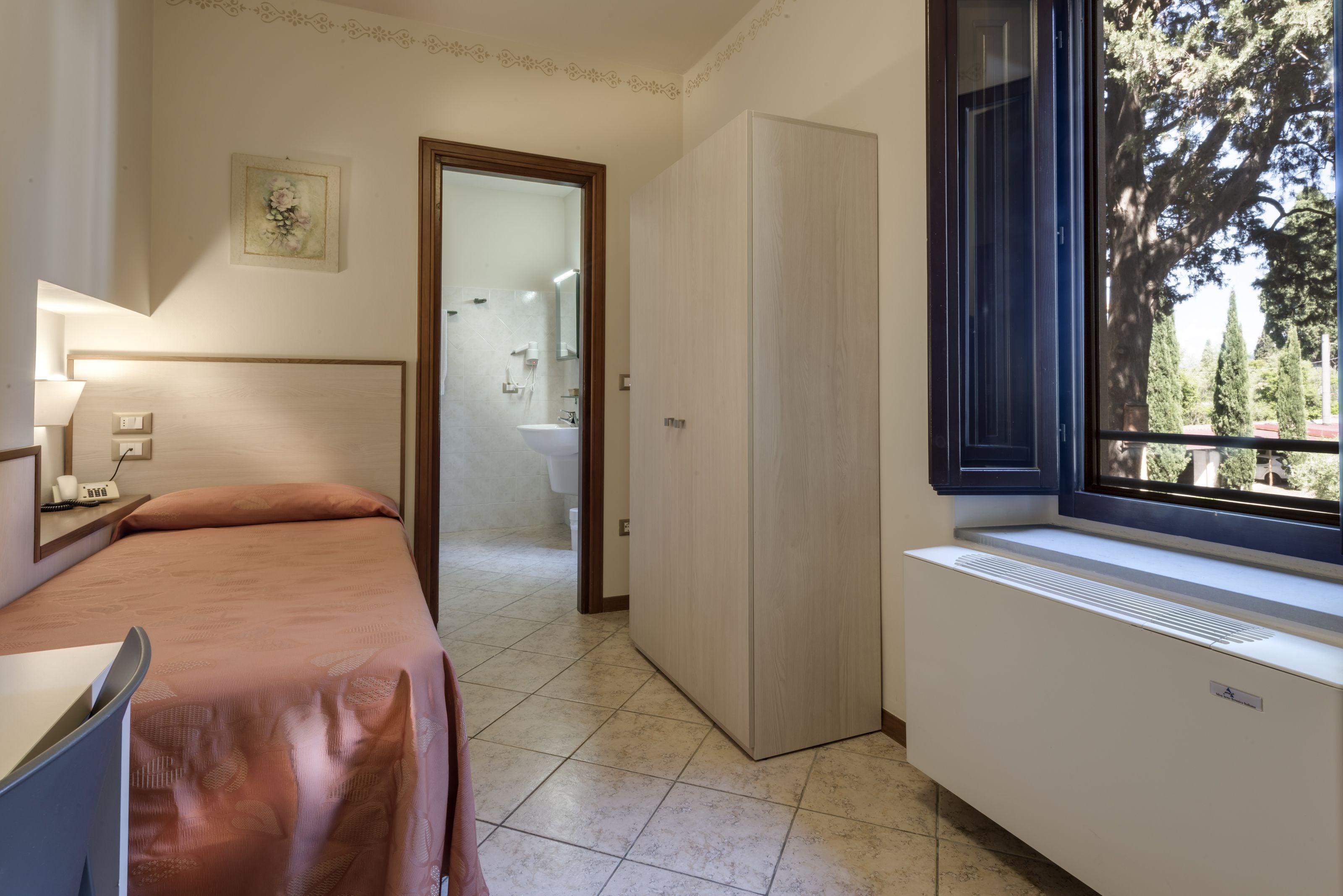 A bedroom with a bed, a desk, a radiator, and a window with a view of trees.