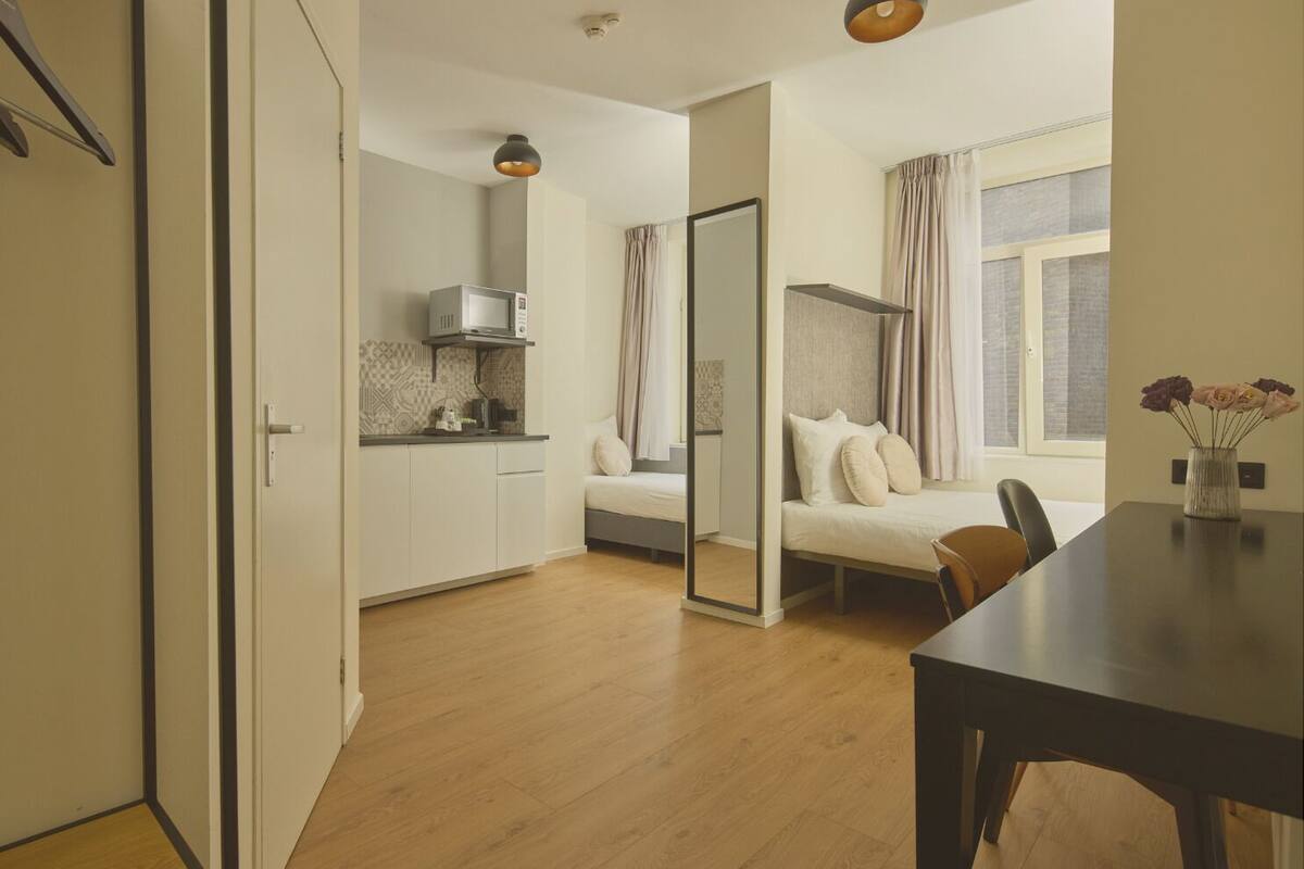 A compact hotel room with a kitchenette, a bed with a headboard, a dining table, and a window with curtains.