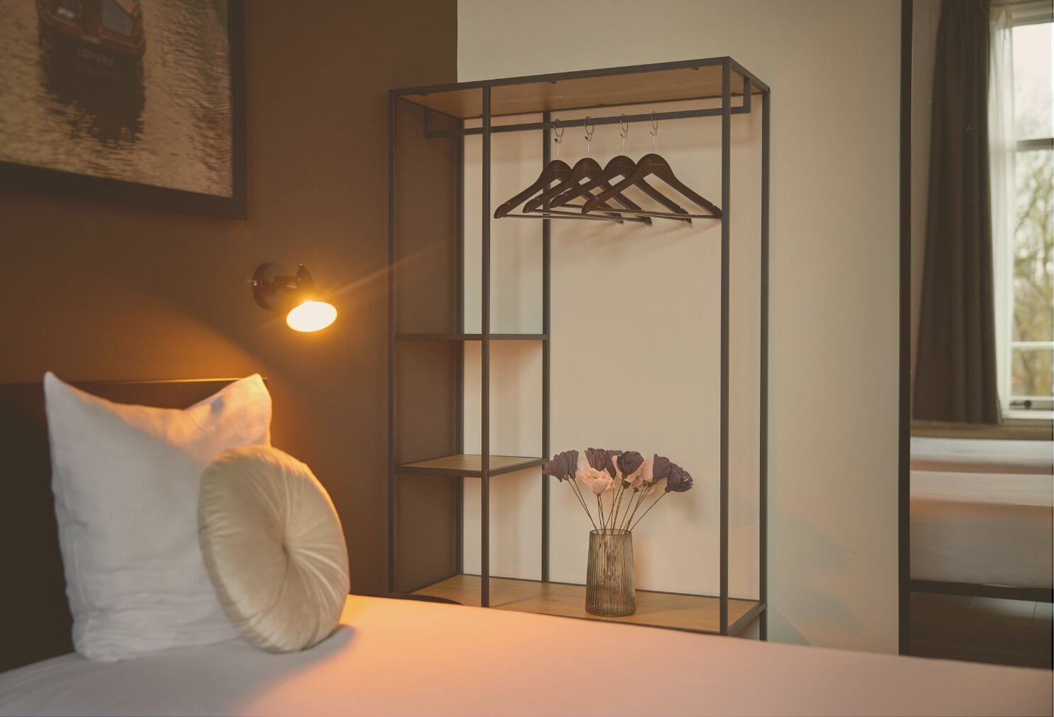 A hotel room with a bed, a wall-mounted shelf with hangers, and a vase with flowers.
