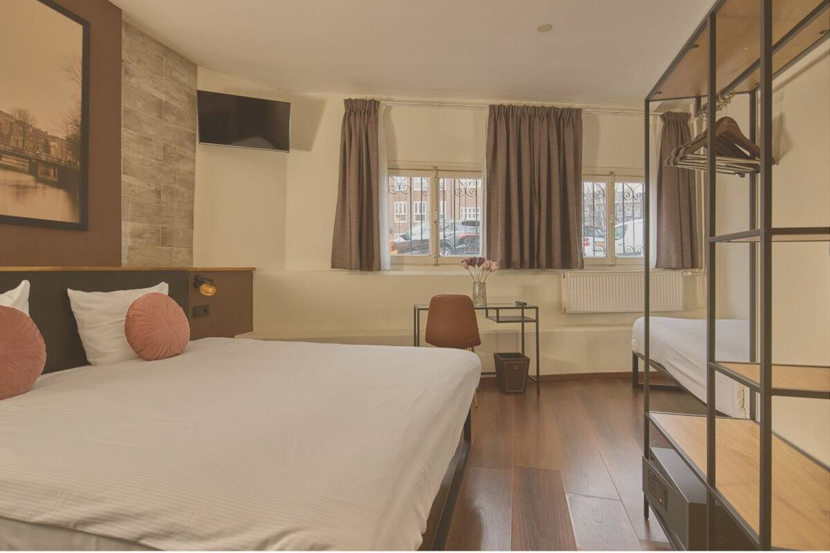 A hotel room with a large bed, a desk, a chair, a radiator, and a window with curtains.