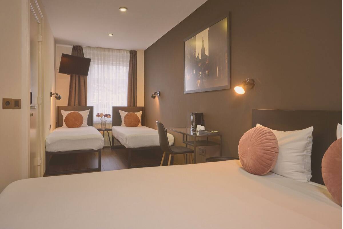 A hotel room with two single beds, a large bed, a desk with a chair, a coffee machine, and a cityscape painting on the wall.