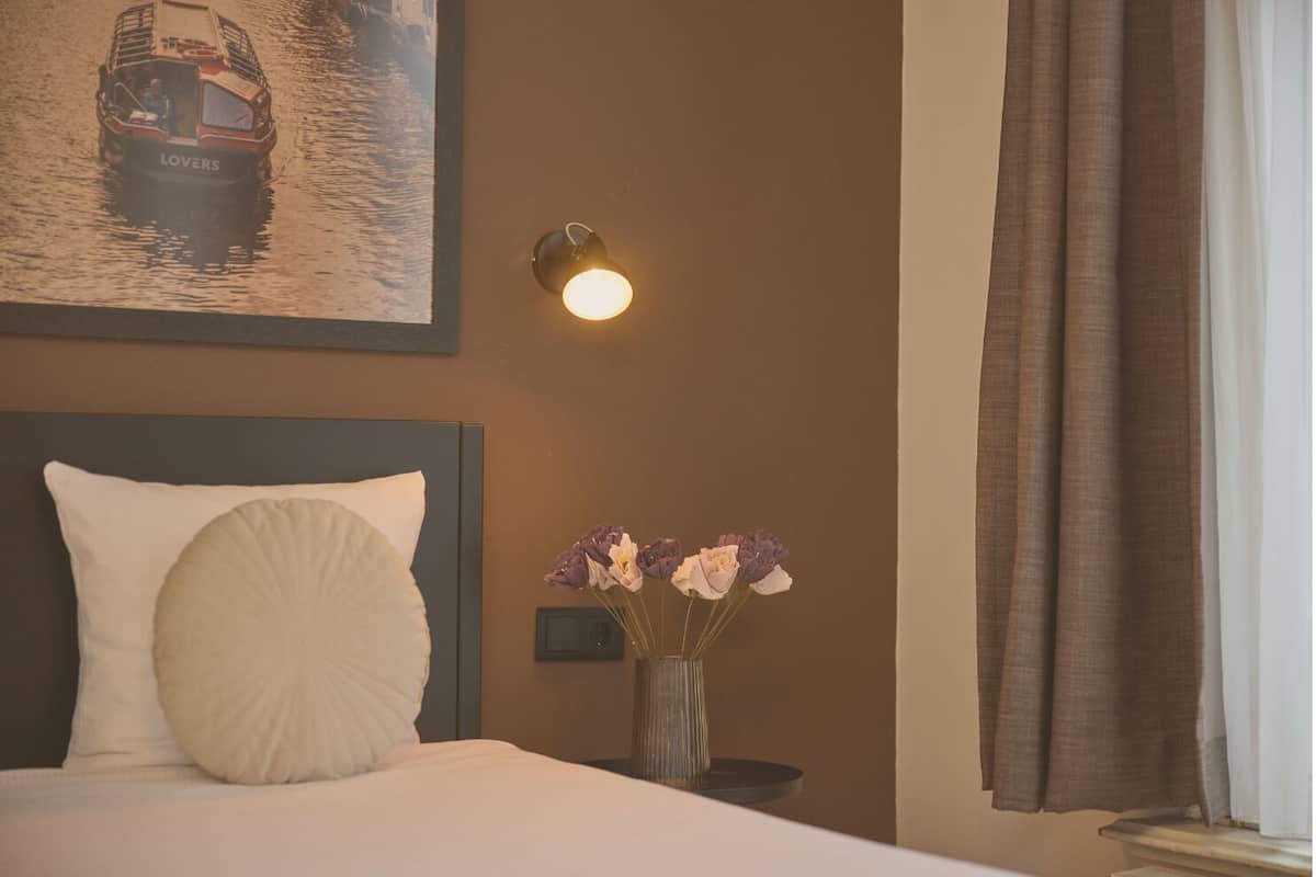A hotel room with a bed, a nightstand with a vase of flowers, a wall-mounted lamp, and a framed picture of a boat on the wall.